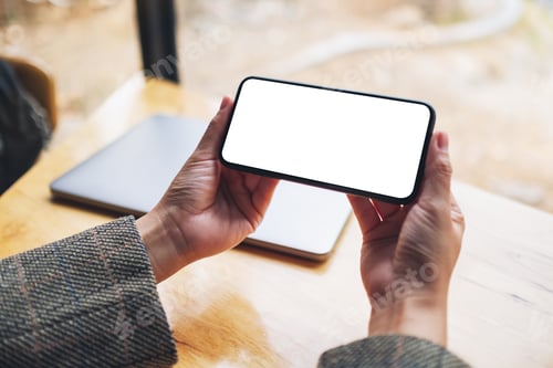 Preview: Mockup image of a woman holding mobile phone with blank white desktop screen with laptop on table