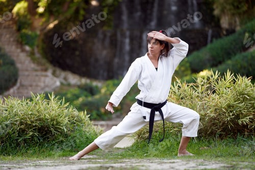 Preview: Woman in white kimono with black belt practicing karate outdoors in nature.