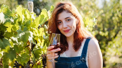 Vista previa: Hermosa chica pelirroja sonríe con una copa de vino entre viñedos en el campo