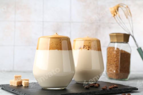 Preview: Delicious dalgona coffee in glasses, brown sugar, beans, granules and whisk on table, closeup