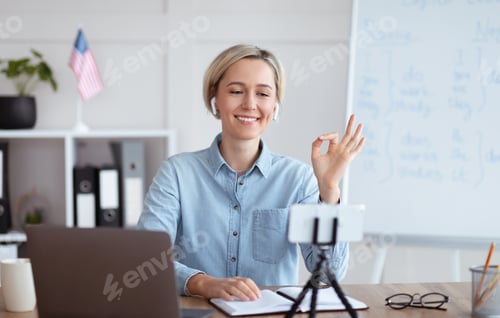 Preview: Positive female teacher giving online English lesson on smartphone, showing OK gesture at home
