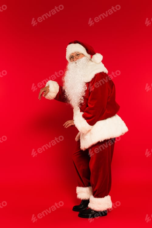 Preview: Full length of bearded santa claus dancing and looking at camera on red background