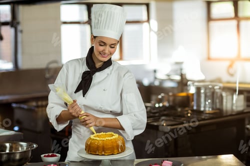 Preview: Female pastry chef piping icing onto bundt cake in commercial kitchen with pastry bag, raspberries