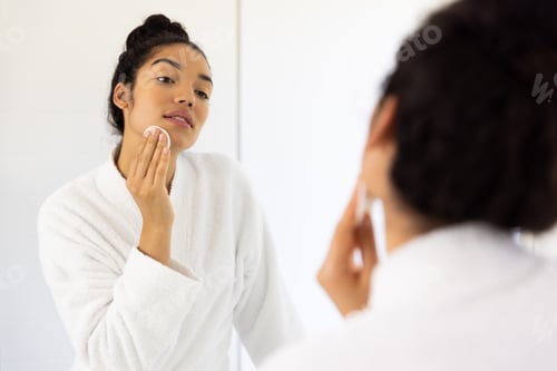Preview: Biracial woman in bathrobe cleansing her face in sunny bathroom