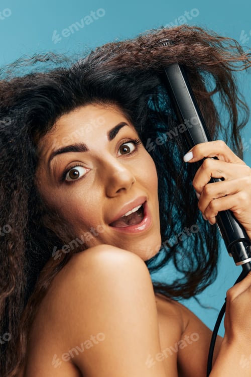 Preview: Excited pretty Latin curly woman using hair straightener, looking aside, posing isolated on blue