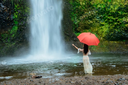 Preview: Woman with red umbrella stands by a waterfall surrounded by lush greenery. Silver Falls, Oregon, USA