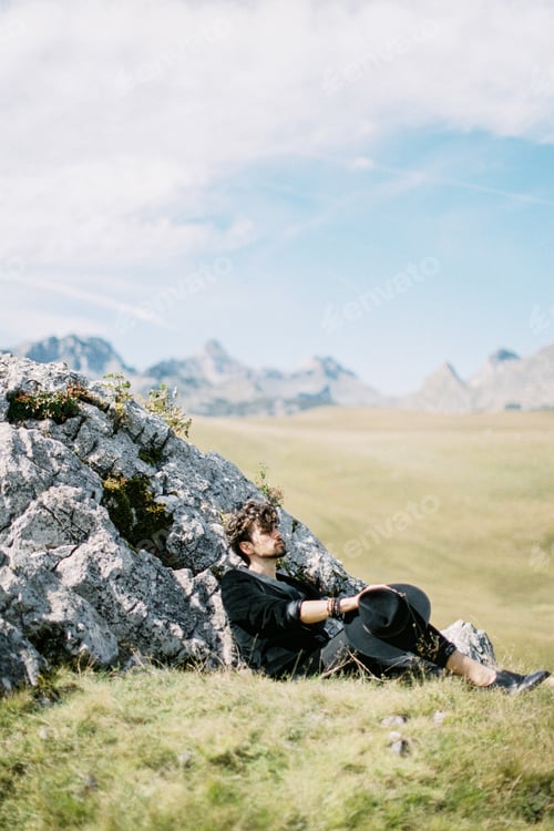 Preview: Young man in a jacket sits on the lawn with his back against a stone
