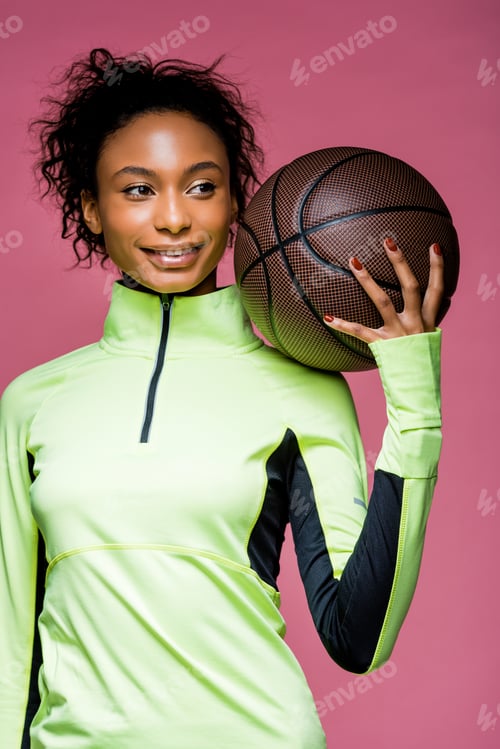 Preview: beautiful smiling african american sportswoman holding basketball isolated on pink