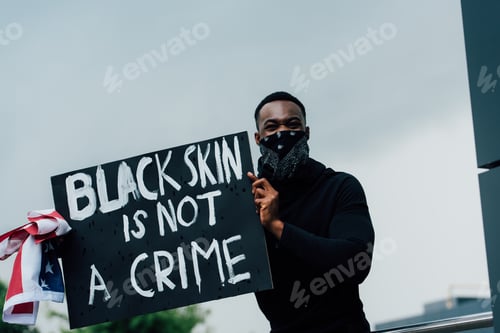 Preview: african american man holding placard with black skin is not a crime lettering and flag of america