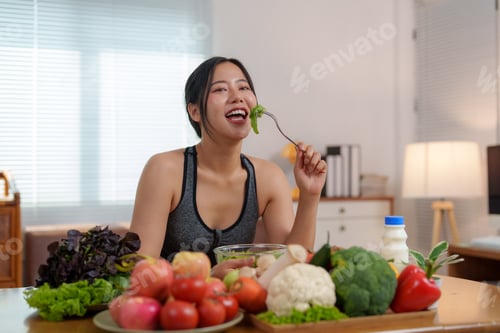 Preview: Happy sportswoman eating healthy salad at home with fresh vegetables on table