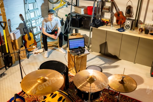 Preview: Happy young restful man sitting in front of drumset, laptop and microphone