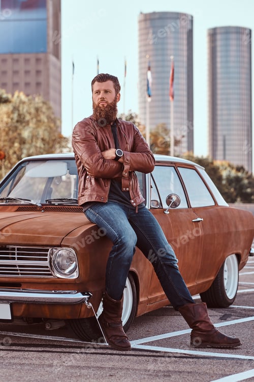 Preview: Bearded male dressed in brown leather jacket and boots with tuned retro car