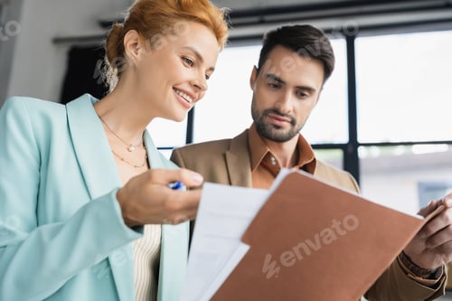 Preview: cheerful redhead businesswoman showing documents to bearded colleague in office