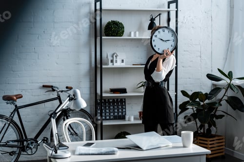 Preview: businesswoman in formal wear holding clock in front of face in modern office