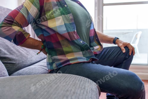 Preview: Older male wincing from lower back discomfort, pressing painful area seated on home living room