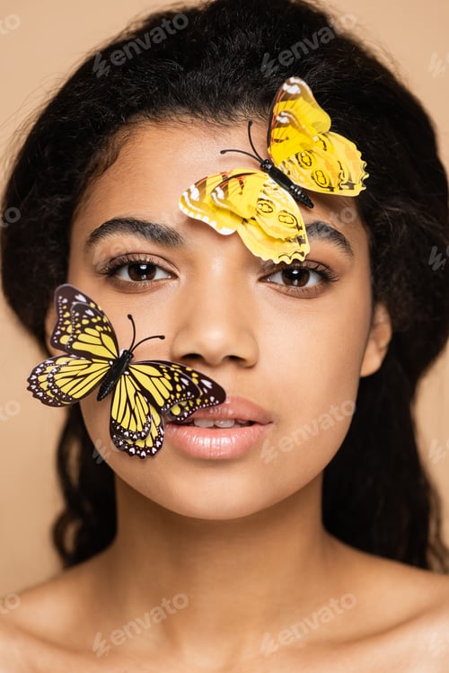 Preview: young african american woman with decorative butterflies on face looking at camera isolated on beige
