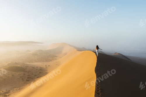 Preview: Woman walking on top of huge sand dune in Morocco Sahara desert. Beautiful warm sun light and mist
