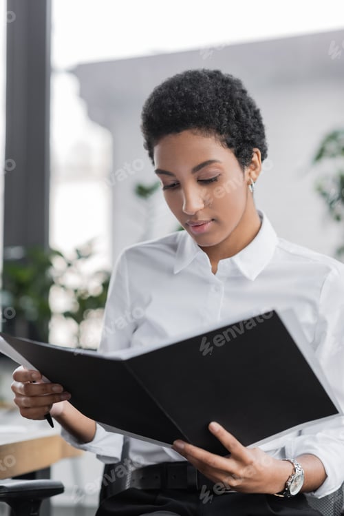 Preview: young african american businesswoman in white blouse reading documents in folder in office