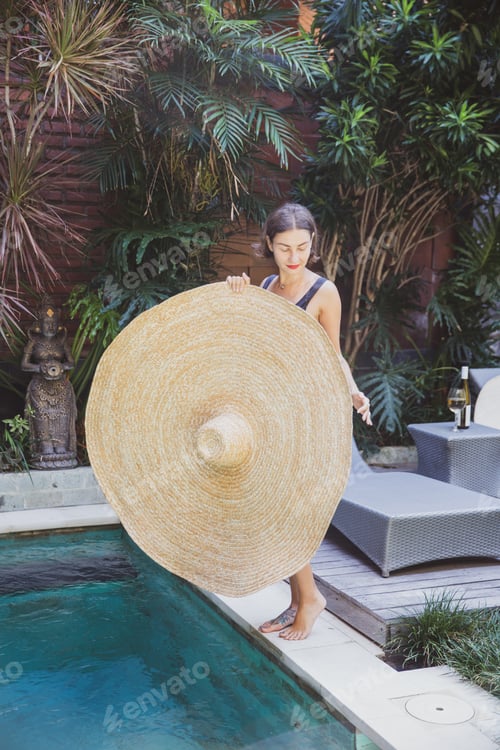 Preview: Beautiful sexy woman holdin big straw hat enjoying vacation by swimming pool and tropical palms