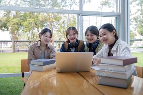 Preview: University students sitting together at table with book and laptop. Happy young people doing group