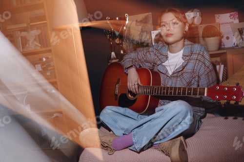 Preview: Portrait of Caucasian Teenager Girl Playing Guitar While Sitting on Bed Indoors