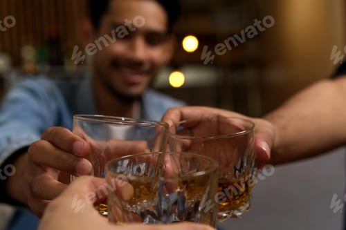 Preview: Close up of glasses clinking between group of friends drinking whiskey at night party in bar.