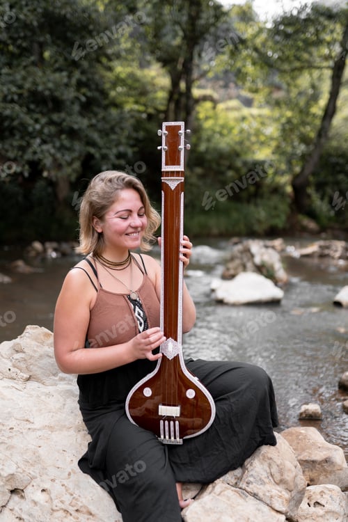 Preview: Portrait of a young adult woman smiling with her indian tanpura sitting on a river rock