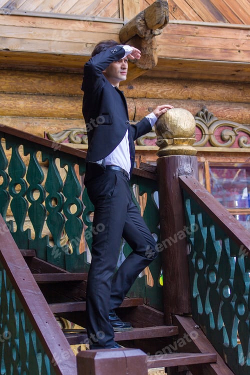 Preview: A young man in an evening suit looks into the distance from a wooden staircase.
