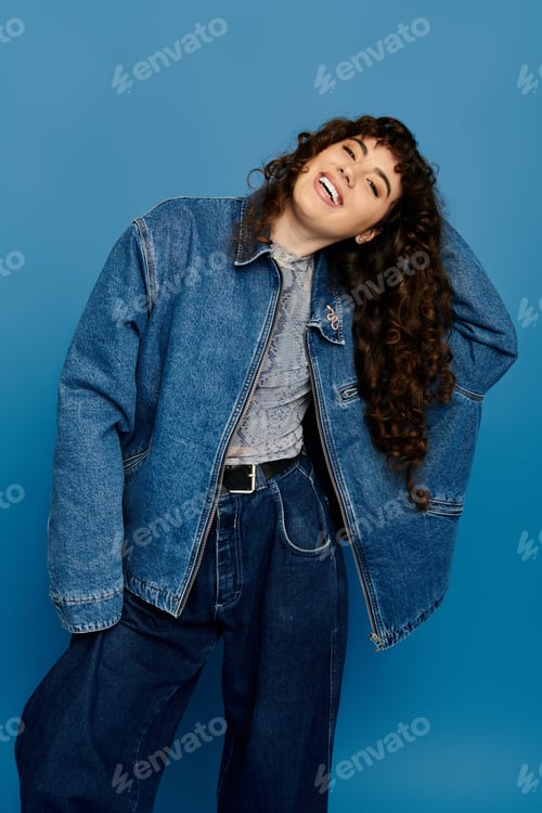 Preview: Stylish young woman expressing joy in trendy denim attire against a vibrant blue backdrop