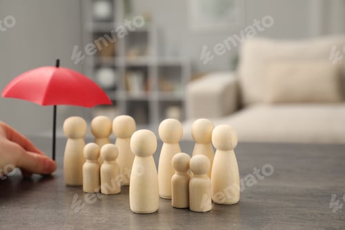 Preview: Woman holding umbrella over human figures at grey table indoors, closeup. Insurance concept