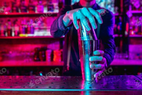 Preview: Close up of barman finishes preparation of alcoholic cocktail with shaker in multicolored neon light
