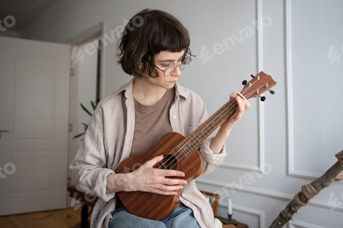 Preview: Focused young woman in glasses learning ukulele chords at home, intent on musical progress