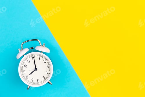 Preview: White alarm clock on a blue and yellow background, flat lay.