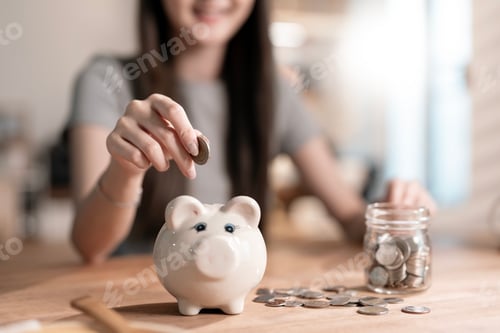 Preview: asian young business woman hand putting coin into piggy bank while sitting at home