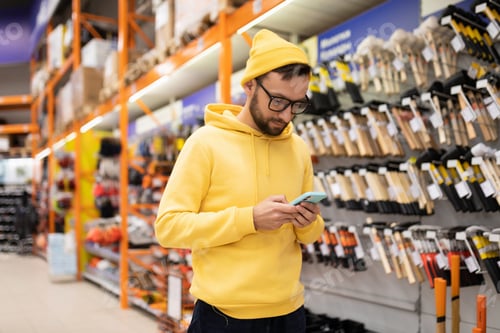 Preview: a young man with a phone in his hands in a hardware store next to a showcase with hammers