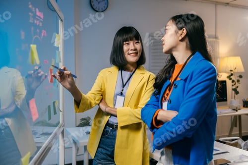 Preview: Two young businesswomen smiling and having a discussion while using a whiteboard during a brainstorm