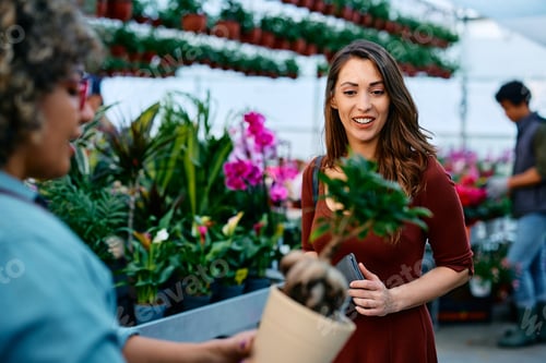 Preview: Happy woman talking to florist while buying pot flowers in garden center.