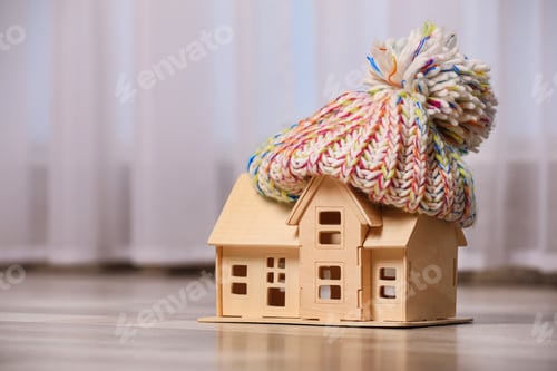 Preview: Wooden house model in hat on floor indoors, space for text. Heating efficiency
