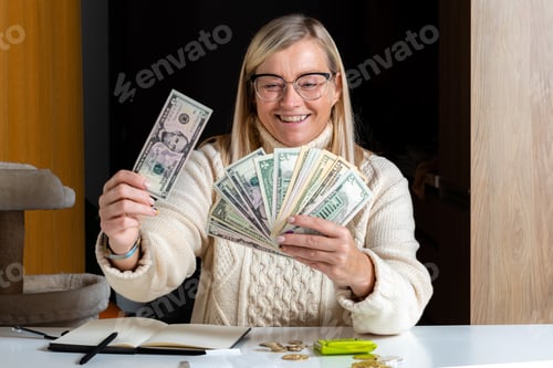 Preview: surprised woman sitting at a table and looking at dollar bills and enjoying the money earned