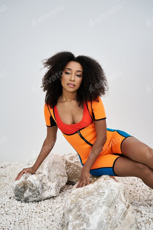 Preview: Stylish young African American woman poses elegantly on textured stones in vibrant attire