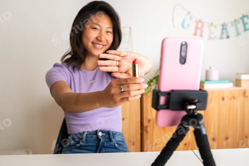 Preview: Happy asian woman showing lipstick on camera, live streaming make up tutorial using phone on tripod.