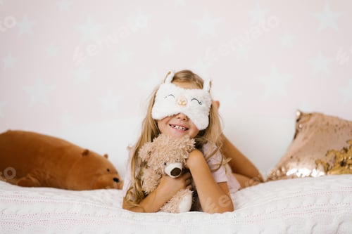 Preview: Cute Little Girl Hugging Teddy Bear is Lying on Cozy Bed, with Sleeping Mask and Smiling