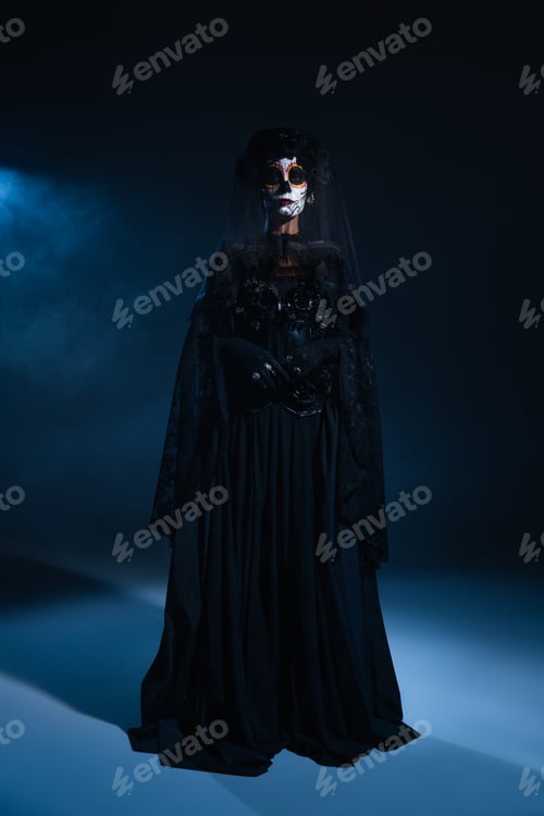 Preview: full length of woman in traditional santa muerte makeup and black dress standing in blue light