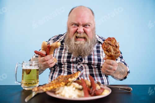 Preview: Angry man with overweight holds chicken leg and sausages at table with greasy food