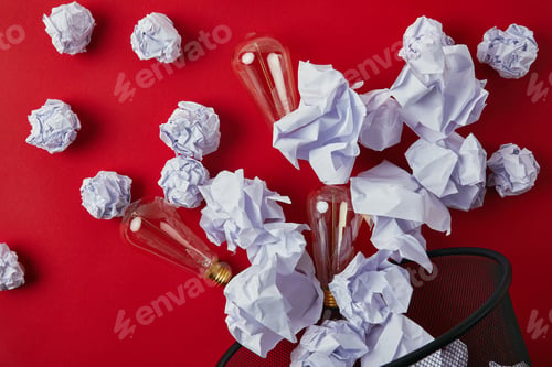 Preview: top view of crumpled papers with vintage incandescent lamps spilled from trash bin on red tabletop