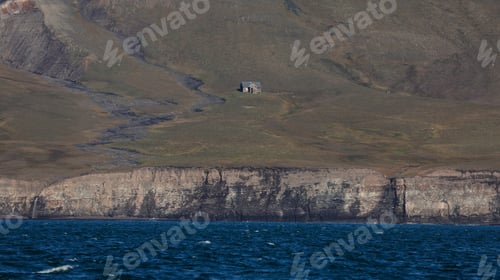 Preview: A small building stands alone on the sloping hillside of Svalbard overlooking the blue waters
