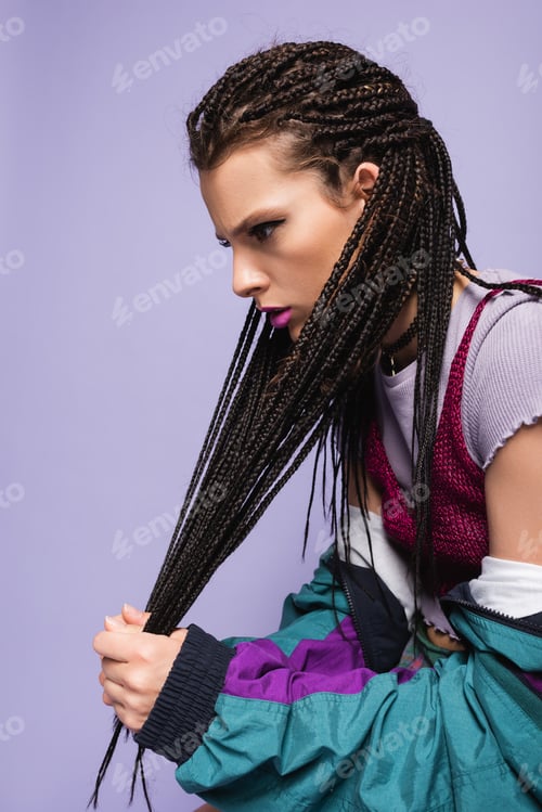 Preview: young woman in vintage clothes touching braided dreadlocks isolated on purple