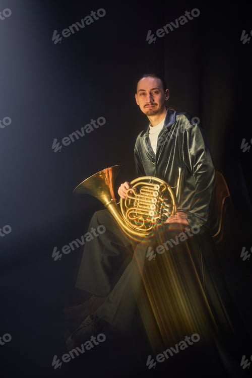 Preview: Portrait of Male Musician Posing with French Horn Sitting in Dimly Lit Studio against Black