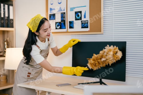 Preview: Young smiling maid wearing yellow gloves is cleaning a computer screen with a feather duster in an o