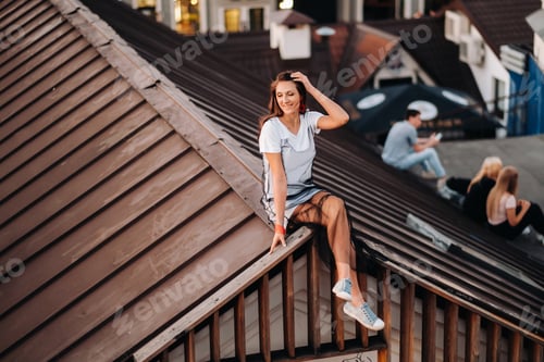 Preview: A girl sits on the roof of a house in the city in the evening. Portrait of a model in a dress and